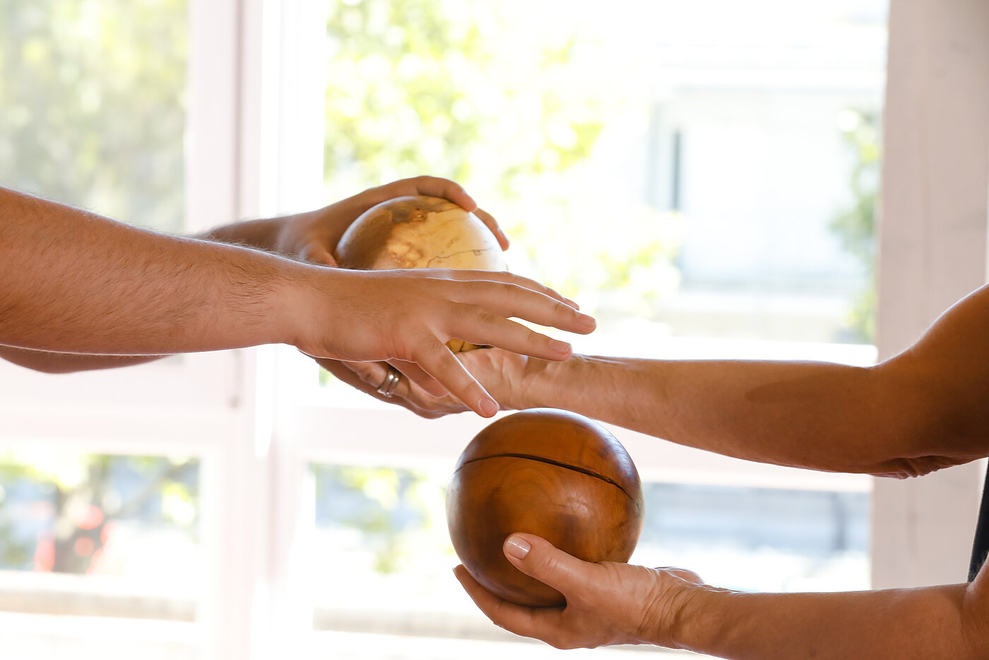 Zwei Personen stehen sich gegenüber. Eine Person übergibt eine Holzkugel mit ihrer Hand an die andere Person, die die Kugel mit ihrer Hand annimmt.